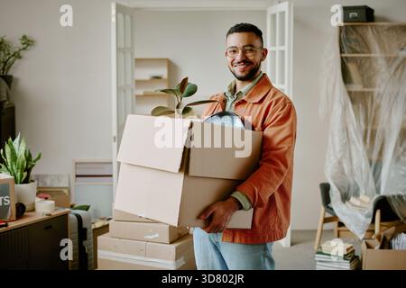 Giovane adulto che sorride mentre trasporta scatole in movimento durante i lavori di ristrutturazione Foto Stock