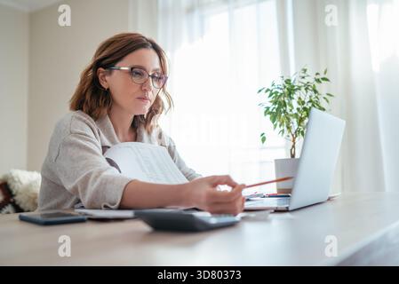 Bella donna di mezza età in bicchieri che contano il budget aziendale di famiglia guardando le bollette mensili e i documenti in casa soggiorno ufficio. Piccole imprese Foto Stock