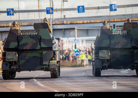 Due veicoli corazzati NATO che si muovono lungo una strada urbana con soldati visibili sulle piattaforme posteriori e spettatori che seguono il percorso durante una def pubblica Foto Stock