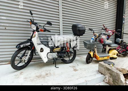 Fila di ciclomotori parcheggiati e scooter elettrici di fronte a un otturatore metallico chiuso su un marciapiede in cemento in un'area urbana di kuala lumpur, malesia Foto Stock