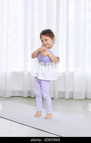 La bambina abbraccia la palla pilates viola, in piedi su un tappetino da yoga in una stanza luminosa Foto Stock