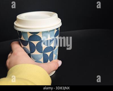 Primo piano di una mano che regge una tazza di caffè da asporto con un motivo geometrico su sfondo scuro Foto Stock