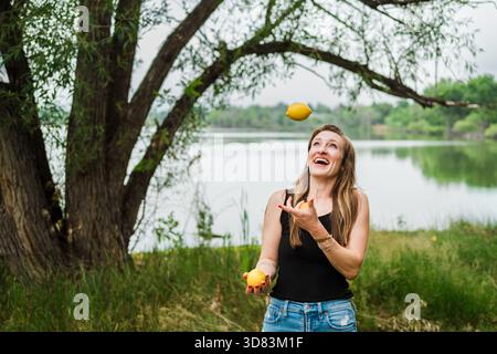 Donna gioiosa che giocherà con i limoni vicino a un lago sotto gli alberi Foto Stock