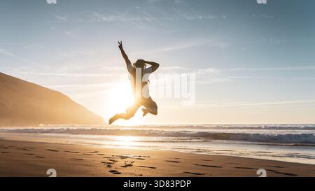 Uomo felice con zaino che salta con le braccia sollevate sulla spiaggia al tramonto - affascinante turista che si gode una vacanza estiva al mare - Traveler Lifestyle An Foto Stock