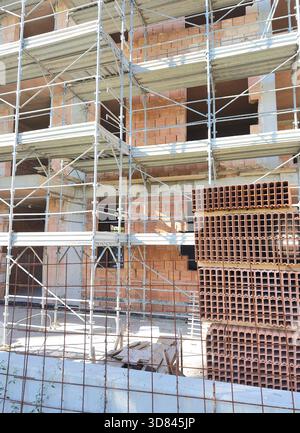 Primo piano di un edificio a più piani con mattoni cavi rossi e ampie impalcature in metallo. Pile di materiali da costruzione in primo piano. Foto Stock