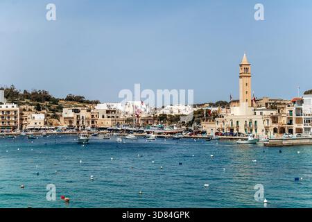 Vista panoramica sul lungomare di Marsaskala con edifici colorati, barche e acque turchesi limpide in un giorno di primavera soleggiato, Malta, Europa Foto Stock