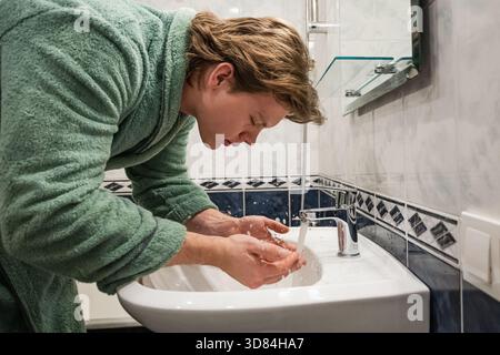Giovane in accappatoio lavarsi il viso con acqua sopra il lavandino del bagno come parte della sua routine di igiene mattutina e cura della pelle. Vista laterale. Foto Stock