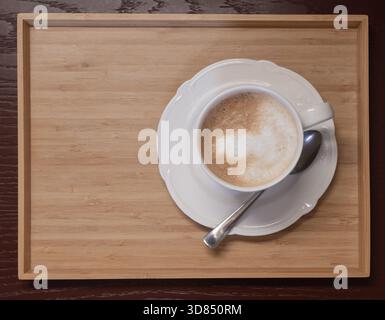 Cappuccino con schiuma cremosa servito in una tazza bianca su un vassoio di legno con cucchiaio. Ambiente accogliente e caldo per caffe', colazione e temi di stile di vita. Foto Stock