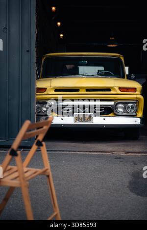 Il classico pick-up GMC giallo brillante degli anni '1960 sbircia fuori dalla porta di un garage buio. Una sedia pieghevole in legno è sfocata in primo piano. Foto Stock