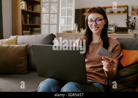 Giovane donna caucasica che usa il laptop e tiene in mano la carta di credito mentre fa shopping online Foto Stock