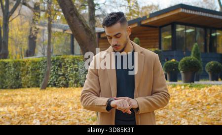 Preoccupato ispanico ragazzo nel parco autunnale guardarsi intorno in attesa di incontro guardando guardare al polso deluso nervoso attesa triste uomo in piedi all'aperto Foto Stock