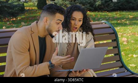 Felice coppia ispanica emotiva che vince con un laptop nel parco autunnale della città seduto sulla panchina famiglia uomo arabo donna caucasica ragazza sposata multirazziale Foto Stock