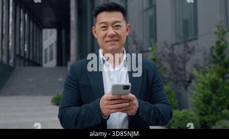 Uomo d'affari maturo asiatico con smartphone uomo d'affari coreano con cellulare sorridente guardando la macchina fotografica in città felice giapponese lavoratore maschile cinese Foto Stock