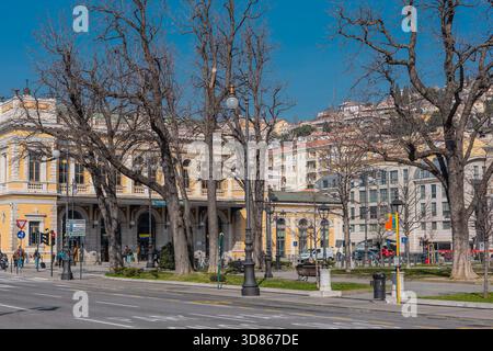 Una vista soleggiata su Piazza della Libert di Trieste dominata dalla grandiosa facciata gialla della stazione ferroviaria di Trieste centrale, con alberi e pedoni. Foto Stock