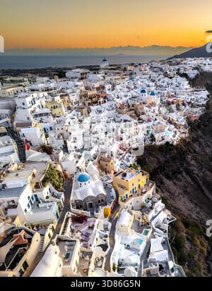 Veduta aerea degli edifici bianchi che scendono lungo la scogliera verso il mare turchese, dove le chiese dalla cupola blu si distinguono contro il caldo tramonto Foto Stock