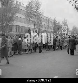 Slavyansk, Ucraina, URSS - 1970s: Una sfilata di manifestazioni festive in via Karl Marx (ora via Tsentralna). Le colonne dei residenti marciano oltre l'edificio modernista "Dom Byta" (House of Life Services). I partecipanti portano bandiere, palloncini, striscioni con slogan come "abbiamo bisogno di pace!" e ritratti di leader sovietici, tra cui Leonid Brezhnev. La gente è vestita con cappotti autunnali. Questa foto d'archivio in bianco e nero cattura i rituali ideologici, la moda e l'atmosfera sociale della tranquilla città industriale di Donbas. Foto Stock