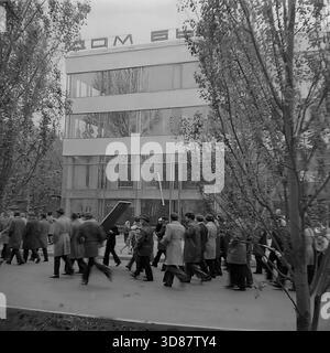 Slavyansk, Ucraina, URSS - 1970s: Una sfilata di manifestazioni festive in via Karl Marx (ora via Tsentralna). Le colonne dei residenti marciano oltre l'edificio modernista "Dom Byta" (House of Life Services). I partecipanti portano bandiere, palloncini, striscioni con slogan come "abbiamo bisogno di pace!" e ritratti di leader sovietici, tra cui Leonid Brezhnev. La gente è vestita con cappotti autunnali. Questa foto d'archivio in bianco e nero cattura i rituali ideologici, la moda e l'atmosfera sociale della tranquilla città industriale di Donbas. Foto Stock
