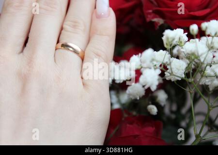 Una mano con un lucente anello d'oro è graziosamente posizionata vicino a un mazzo di rose rosse e fiori bianchi. Questa scena cattura un'atmosfera romantica e intima Foto Stock