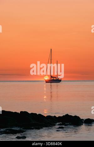 Guarda il mare con una barca a vela che si staglia contro l'alba o il tramonto arancione, visto da dietro le scure rocce costiere Foto Stock