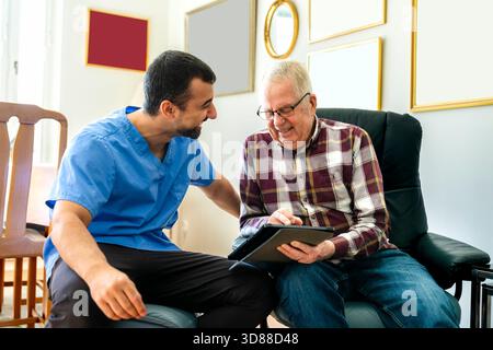 Un operatore sanitario maschile felice seduto con un anziano che utilizza un tablet PC a casa Foto Stock