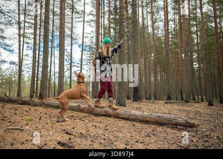 Donna gioiosa che si bilancia attentamente sul tronco dell'albero caduto, il cane attento osserva il proprietario e il salto. Giocosa escursionista femminile nel bosco divertiti con il cucciolo, navigando tra i tronchi della foresta con curiosi canini || modello rilasciato Foto Stock
