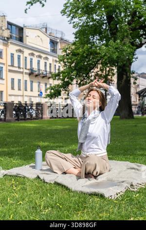 Donna rilassata che allunga le braccia verso l'alto e riposa su una coperta nell'erba del parco, fuggendo dallo stress nelle calde estate. Donna che medita sotto il sole con una bottiglia d'acqua, abbronzatura e rinfresco, rigenerarsi nella natura urbana || modello rilasciato Foto Stock