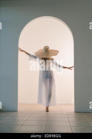 Questa immagine mostra una donna con una veste bianca e un cappello di paglia che cammina attraverso un grande arco bianco. I capelli delle donne sono biondi e ha le braccia estese ai lati mentre cammina attraverso la porta. La luce è morbida e diffusa, creando un atmo da sogno || modello rilasciato Foto Stock