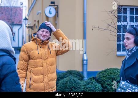 Tre persone stanno conversando all'aperto, vestite con caldo abbigliamento invernale, con una parete gialla luminosa e lussureggiante vegetazione che crea un'atmosfera allegra Foto Stock
