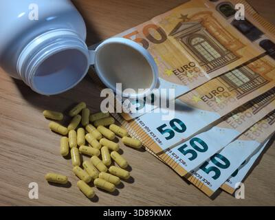 Costo dei farmaci: Pillole e euro - pillole gialle fuoriescono da una bottiglia bianca accanto a banconote da 50 euro su un tavolo di legno, simboleggiando l'alta media Foto Stock