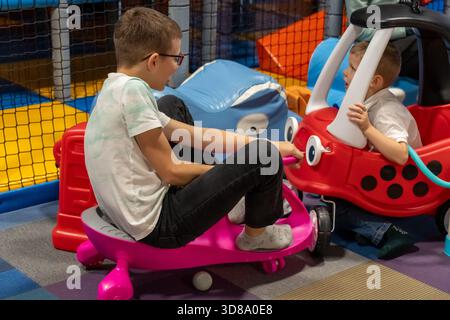 Due fratelli che cavalcano macchinine colorate nel parco giochi al coperto con un bambino più grande su un vagone e più giovane in un'accogliente coupé. Divertimento per i fratelli, ingresso al chiuso Foto Stock