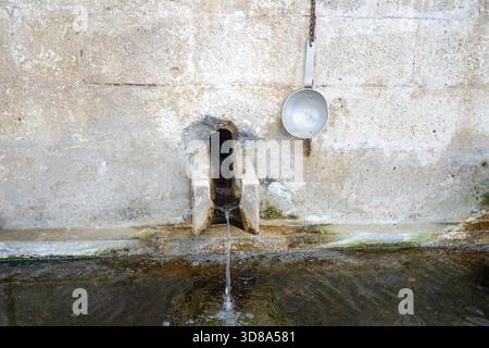 Una semplice e rustica fontana a muro in pietra con un singolo beccuccio d'acqua che alimenta una vaschetta, con un mestolo metallico attaccato per bere, nel villaggio di Melampes. Foto Stock
