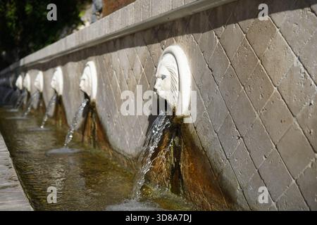 Vista laterale di diversi beccucci di testa di leone lungo la fontana di Kefalovrisi a Spili. Il lungo condotto cattura l'acqua corrente sotto la luce solare intensa. Foto Stock