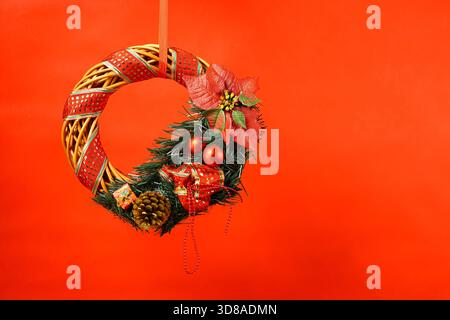 Tradizionale corona natalizia decorata con vegetazione di pino, nastri rossi, baubles, pinne e poinsettia, appesi su uno sfondo rosso brillante Foto Stock