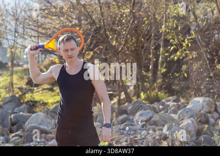 L'atletico con canotta nera regala una vivace racchetta sulla spalla e la palla in mano, in piedi su terreni rocciosi con alberi sullo sfondo. Foto Stock