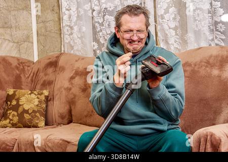 Spazzola aspirapolvere, uomo di mezza età si concentra sulla rimozione dello sporco dagli utensili domestici, l'espressione del viso mostra un chiaro disgusto durante le attività più semplici. Foto Stock