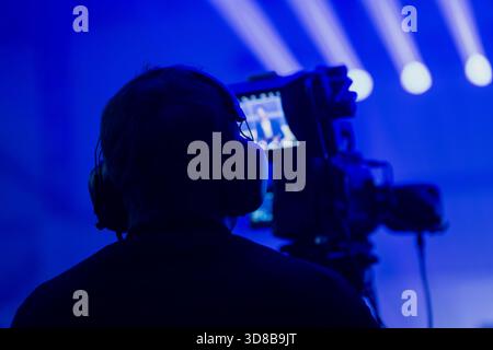 Videografo sagomato che utilizza una fotocamera professionale durante uno spettacolo illuminato in blu con forti fasci di luce che dimostrano i moderni media e la produzione di eventi Foto Stock