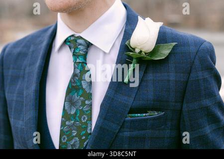 Un uomo ben vestito presenta un abito blu con una cravatta floreale e una rosa bianca nel suo risvolto in un elegante raduno all'aperto. Foto Stock