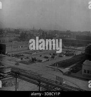 Slavyansk, RSS Ucraina, Unione Sovietica. 1970. Vista panoramica ad angolo alto del cantiere logistico presso l'impianto di isolamento delle armature (AIZ). In primo piano, enormi tubi di riscaldamento isolati attraversano il telaio. Il vasto territorio lastricato è pieno di pile di prodotti ceramici e casse di legno. I lavoratori sono impegnati a caricare merci su carrelli e vagoni ferroviari vicino al magazzino. Un paesaggio industriale impressionante con cisterne fumé. La vita nel pacifico Donbas prima della guerra. Foto Stock