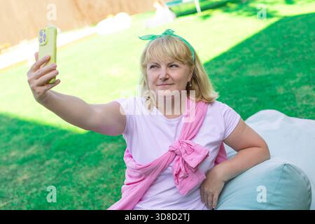 Una donna bionda di mezza età con abiti luminosi si fa un allegro selfie mentre si siede su un prato verde all'aperto. Foto Stock