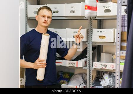 L'uomo guarda la fotocamera, punta il dito verso il pacco su un rack metallico mentre afferra il rotolo di imballaggio nella mano sinistra. Foto Stock