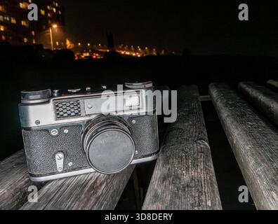 Videocamera vintage su panca in legno di notte con luci urbane sfocate e lampioni che illuminano lo sfondo scuro. Foto Stock