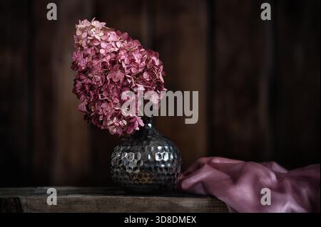 Composizione Still Life vintage con ortensie rosa essiccate in vaso marrone decorativo con motivo testurizzato su una superficie rustica in legno. Foto Stock