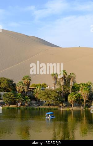 Oasi di Huacachina nel deserto dell'Ica in Perù, con torreggianti dune di sabbia, palme e visitatori che esplorano il paesaggio panoramico del deserto. Foto Stock