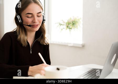 operatore donna con auricolare che scrive appunti su un notebook che lavora da remoto in cucina. copia spazio Foto Stock