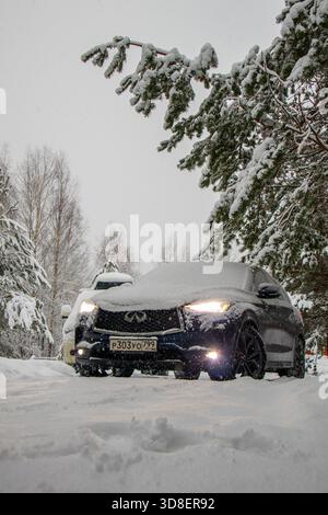 Mosca, Russia - 13 dicembre 2024: Vista frontale ravvicinata di un dark crossover Infiniti QX50, pesantemente spolverato di neve e parcheggiato sotto pino carico di neve Foto Stock