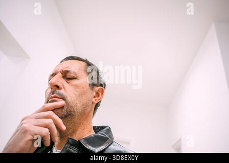 Uomo maturo con giacca in pelle in posizione interna, toccando il mento con un'espressione attenta e contemplativa. Messa a fuoco selettiva. Foto Stock