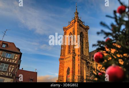 La calda luce del sole illumina la torre gotica della chiesa di San Martino al tramonto, dietro un albero di Natale sfocato con ornamenti rossi a Colmar, in Francia Foto Stock