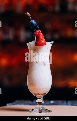 Bevanda cremosa e miscelata servita in un alto vetro curvo con sfumature di cioccolato o caffè, condita da un vivace spiedino di frutti di bosco freschi Foto Stock