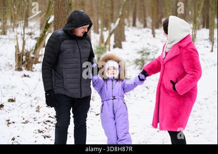 Passeggiata in famiglia attraverso la foresta innevata. Madre, padre e figlia si godono una passeggiata invernale tra alberi innevati, osservando amorevolmente l'un l'altro e tenendosi per mano Foto Stock