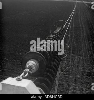 Regione di Donetsk, RSS Ucraina, Unione Sovietica. 1970 anni Una vista mozzafiato ad angolo alto dalla parte superiore di una torre di trasmissione ad alta tensione (750 kV). La telecamera guarda verso il basso lungo enormi doppie corde di isolanti di tensione in vetro che si estendono sui campi agricoli. Piccoli camion e trattori sovietici sono visibili sul terreno, sottolineando l'immensa scala della struttura. Una prospettiva unica che mostra l'ambiente quotidiano degli uomini di linea che costruiscono la rete energetica. La vita nel pacifico Donbas prima della guerra. Foto Stock